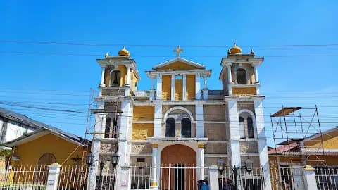 A yellow and white church facade with twin bell towers and scaffolding stands against a clear blue sky in a town center.
