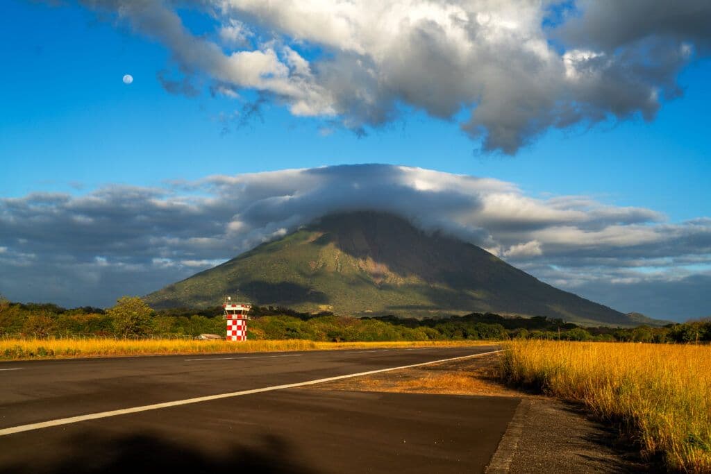 A paved rural road stretches toward a large, cloud-capped volcano under a partly cloudy blue sky.