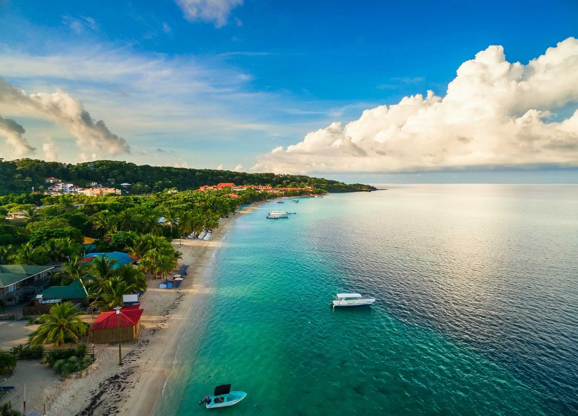 Aerial view of a Caribbean coastline with turquoise water, sandy beach, palm trees, small boats, and coastal buildings under a partly cloudy sky.