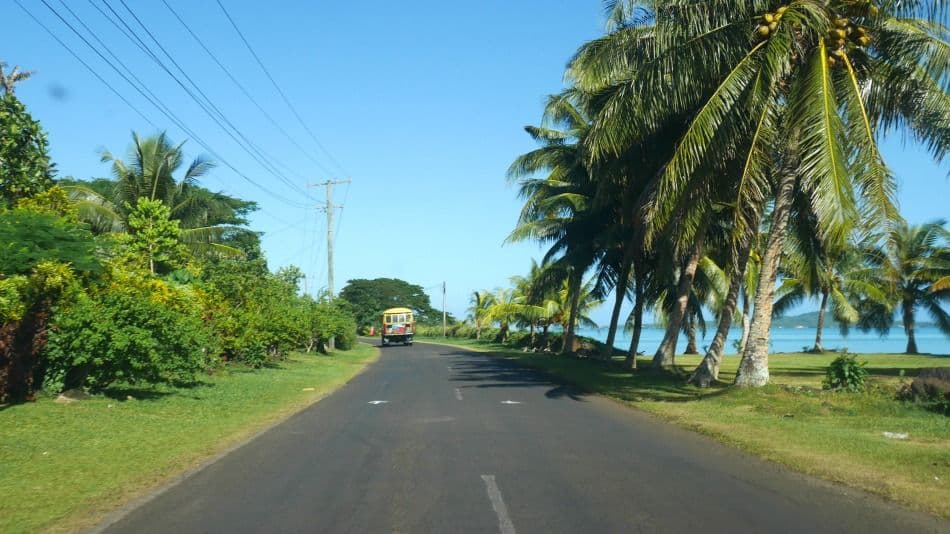 Coastal road with a yellow vehicle, palm trees, and ocean view under a clear blue sky in Samoa.