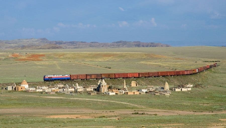 A freight train crosses flat steppe grassland in western Kazakhstan with distant hills and a small settlement in the foreground.