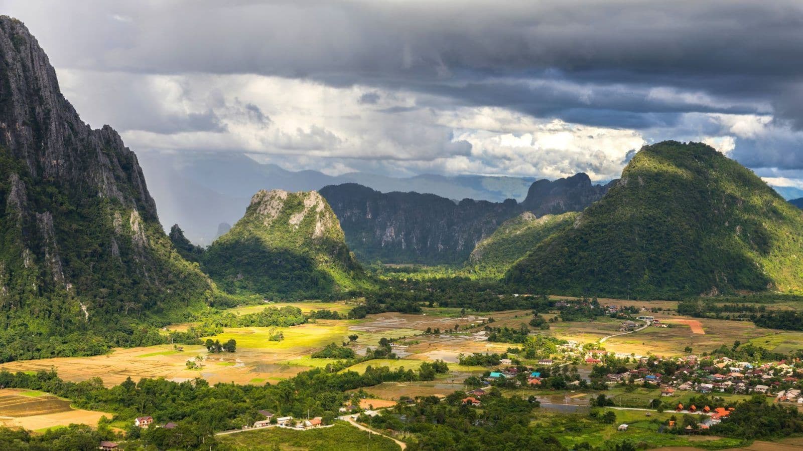 Dramatic landscape featuring limestone karsts and lush green valleys under a cloudy sky, with scattered villages and agricultural fields in the foreground.
