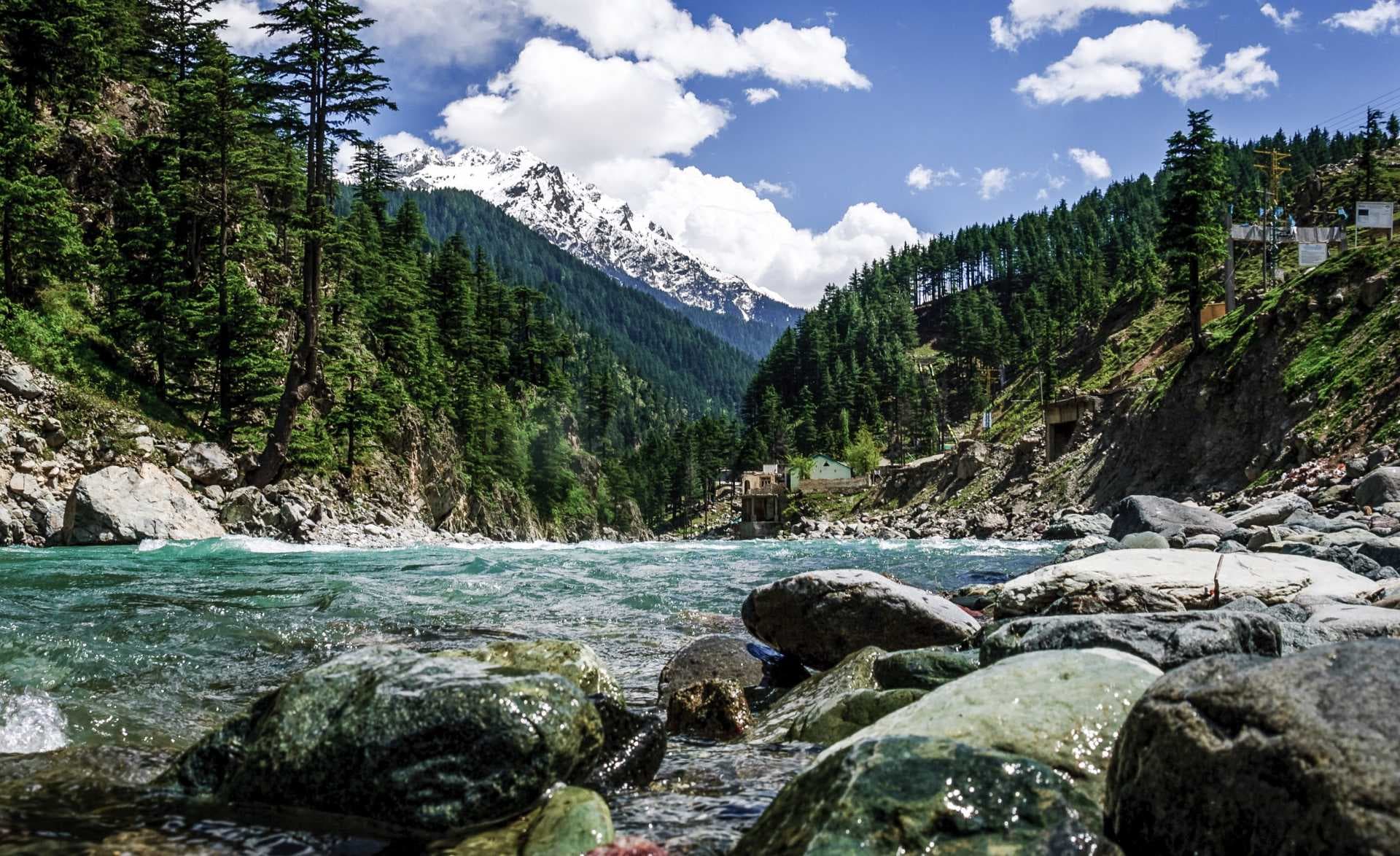 Turquoise river flows through a forested mountain valley with snow-capped peaks and a small settlement visible in the distance.
