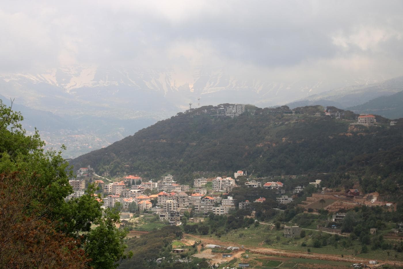 A scenic landscape view of a hillside town with terracotta-roofed buildings, lush greenery, and snow-capped mountains in the hazy distance.